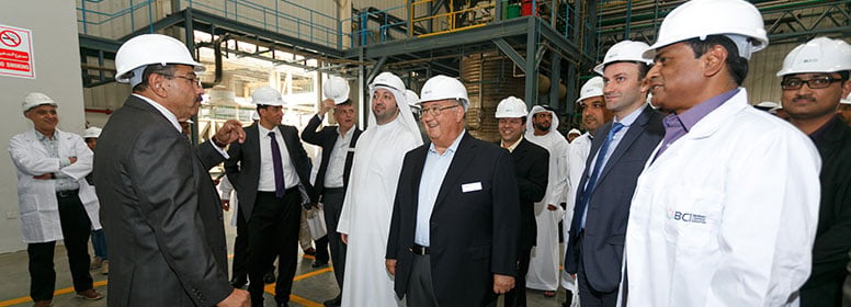 A group of men wearing white hard hats, including individuals in suits and white lab coats, touring the interior of an industrial facility.