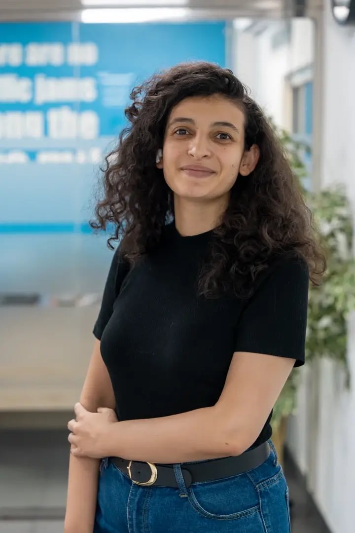 Young woman with curly hair wearing black top and jeans smiling in modern office with blue signage