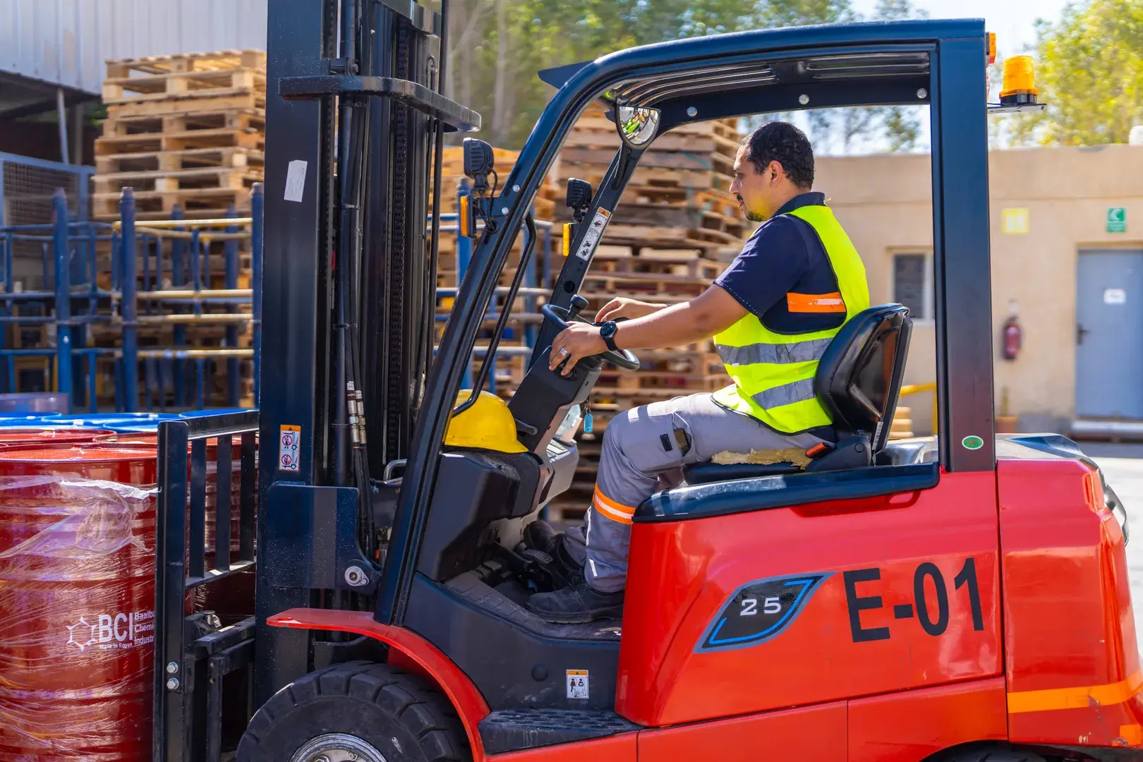 Forklift operator in safety vest driving red forklift E 01 at warehouse with wooden pallets in background