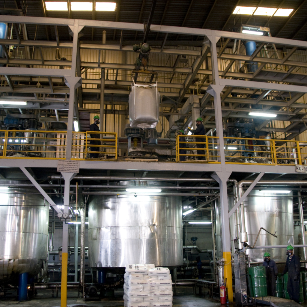 An industrial factory interior featuring large stainless steel tanks on the ground level and workers wearing hard hats on a metal mezzanine overlooking a suspended bulk bag